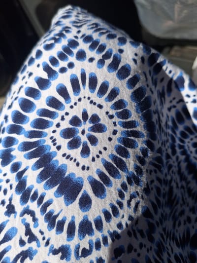 This is a close-up, high-angle shot of a white fabric with a repeating pattern of blue, petal-like shapes and dots. The fabric appears to be slightly creased and is illuminated by sunlight, casting a shadow across part of the pattern. The pattern resembles stylized flowers or perhaps an abstract representation of a natural motif. The texture of the fabric, a woven material, is visible due to the close proximity of the camera. There are no people or clear objects visible, and the setting is abstract, focusing entirely on the textile. The lighting suggests it could be daytime, possibly outdoors or near a window. No text is visible in the image.
