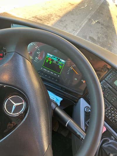 The image depicts the interior of a vehicle, specifically the driver's side. A black Mercedes-Benz steering wheel dominates the foreground, featuring the iconic Mercedes-Benz logo. Behind the steering wheel is a digital dashboard displaying various vehicle information, including speed in km/h, engine RPM, outside temperature at 27°C, and system information like "CONNECT" and the time "20:13." Various buttons such as "SYSTEM," "QUIT," "RESET," and "INFO" are present below the digital display. Above the dashboard, a radio unit is visible, with buttons and functions labeled "ON," "FM/SCRL," "CCS Professional," "COCKPIT," "RADIO," "CD," "ENTER," "NAVI," "MENU," "AUX," and "CAM." A lever with several vehicle control settings, including lights and limiters, is located to the right of the steering wheel. The dashboard area seems to be slightly messy, with a light blue cloth visible behind the lever. The windshield is visible in the upper left, showing a dirty surface. The location of the image is Dimitrovgrad Municipality, Serbia.