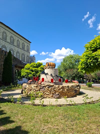 The image depicts a picturesque outdoor scene likely at a winery or similar establishment in Milestii Mici, Moldova, under a clear blue sky with scattered white clouds. In the foreground, a circular, multi-tiered fountain constructed from rough-hewn stone dominates the center. It is adorned with several large, red wine glasses and clay amphorae. Perched atop the fountain is a wooden barrel. Surrounding the fountain are a paved walkway, lush green grass, and young rose bushes. To the left, a building with a stone facade, arched windows, and a tiled roof stands prominently, suggesting a historical or European architectural style. A tall, conical evergreen tree stands next to the building. Behind the fountain, vibrant green trees and foliage create a natural backdrop, with glimpses of parked vehicles and streetlights in the distance, hinting at the edge of the property. The overall atmosphere is serene and suggests a warm, sunny day.