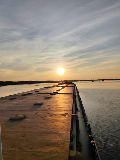 A low-angle, wide shot captures a serene sunset over a body of water, with a long, flat structure dominating the foreground. This structure, possibly a barge or pier, is covered in a weathered, tan material, punctuated by numerous circular, raised hatches. The surface is illuminated by the warm, golden light of the setting sun, which is positioned low in the sky, just above the horizon. Wispy clouds streak across the sky, reflecting the sunset's colors. The water is calm, with gentle ripples catching the light. In the distance, a dark, tree-lined shore is visible, and a few small objects, possibly buoys or distant boats, dot the water. The scene is devoid of people. The location is identified as Shchetinino, Russia.