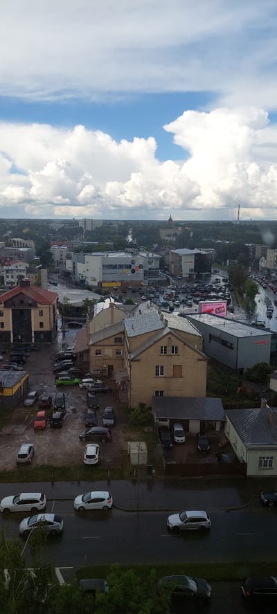 This is an aerial, vertical, full shot of a city in Panevėžys, Lithuania. The sky is filled with dramatic, white cumulus clouds against a bright blue sky. The foreground shows a wet, gray asphalt road with several cars parked and driving on it, indicating it has recently rained or is raining. Several parked cars are visible in a lot to the left, and a larger parking lot is seen further back, packed with vehicles. Buildings are visible throughout the scene, including residential and commercial structures, with one prominent shopping center on the right side of the frame. In the distance, more buildings and trees are seen, with a tall industrial smokestack and a tower in the far background. The overall impression is a somewhat overcast day in an urban environment.