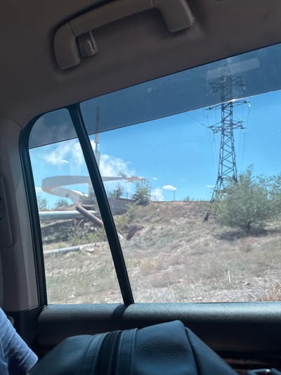 The image, taken from inside a vehicle, shows a view through the rear window. Outside, under a clear blue sky with some clouds, a large power line pylon dominates the right side of the frame. To the left, there's a curved white structure, possibly part of a monument or an architectural feature, with a model airplane positioned in front of it. The landscape appears dry and somewhat barren, with sparse vegetation and a prominent pipe-like structure running horizontally in the middle ground. The car's interior is visible in the foreground, including a gray handle on the ceiling and a dark textured surface of the door panel. The overall scene suggests a location in an industrial or semi-rural area. This imagery is consistent with the area around the monument to the first Soviet cosmonauts in Jezqazğan, Kazakhstan, which features a monument resembling a rocket and an airplane, and is situated in a landscape with industrial infrastructure like power lines and pipelines.