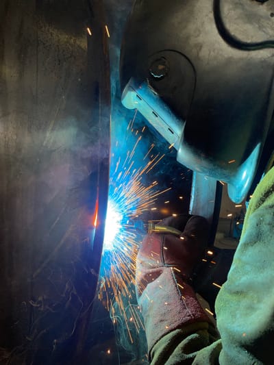 A close-up, medium shot captures a welder in profile, engaged in a welding operation. The welder is wearing a dark welding helmet, a portion of which is visible on the right side of the frame. Protective gear includes a thick, red glove on the left hand, holding a welding torch, and a green, padded sleeve on the right arm. The welding process illuminates the scene with intense blue and white light, accompanied by a shower of bright orange sparks and plumes of white smoke. The background features a large, dark, cylindrical object on the left, likely the workpiece being welded. The environment appears to be an industrial setting. There is no specific indication of the city of Tomba, Montenegro, in this image.