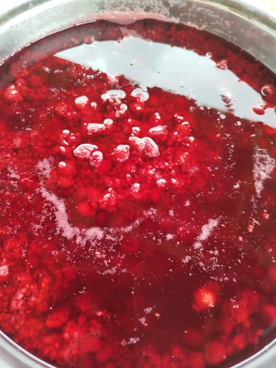 An overhead, close-up shot shows a stainless steel pot filled with bubbling red raspberries and syrup. The raspberries are whole and appear to be cooking down to make jam or preserves. The syrup is a deep, rich red and is actively boiling, with small bubbles rising to the surface. The lighting suggests an indoor setting, likely a kitchen. The image does not contain any information about location within Баравухскі сельскі Савет, Belarus.