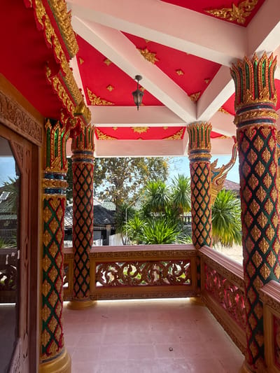 The image depicts an ornate, elevated porch or walkway of a traditional Thai structure located in Baan Tha Po, Thailand. The scene is devoid of human presence or activity.

The ceiling is predominantly red, adorned with gold, intricate patterned motifs, and supported by white structural beams forming a geometric grid. A single dark-colored, ornate lantern hangs centrally from the ceiling. Four richly decorated columns support the roof, featuring vertical bands of gold, green, and red patterns, primarily diamond or stylized floral shapes. The column capitals are elaborate, resembling lotus petals or crowns, in shades of gold and green.

The floor is tiled with light-colored, possibly off-white or light pinkish-brown, square tiles. A decorative balustrade, likely made of wood, with intricate carved scrollwork and floral motifs, runs along the outer edge of the porch, painted in a golden-brown hue. To the far left, a section of a wooden door frame and a reflective glass panel are visible, indicating an entrance to an interior space.

Beyond the balustrade, the outdoor environment reveals lush green tropical foliage, including large palm-like plants and a tree with sparse leaves. In the background, glimpses of other buildings with dark-colored roofs can be seen through the vegetation. The sky is clear blue, indicating bright daylight conditions.