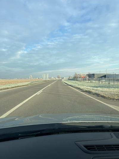 The image depicts a two-lane asphalt road, likely the D37 in Hœrdt, France, viewed from the interior of a vehicle looking forward. The road features a solid white line on the right shoulder and a dashed white centerline. To the left, a brown tilled field is bordered by a frosted grassy verge and a metal guardrail. A triangular warning sign is visible on the left side of the road, followed by a large rectangular sign structure further down. In the distance on the left, residential structures and trees indicate a settlement. To the right, a frosted grassy verge abuts a black metal fence, behind which are dark industrial or commercial buildings with visible light poles and at least two parked white vehicles. A red structure is also discernible within this industrial area. The environment is characterized by widespread white frost covering the ground, indicating cold temperatures. The sky is partially cloudy with patches of blue. No individuals are visible.