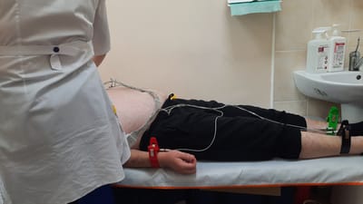 The image shows a male patient lying on a medical examination table, connected to an electrocardiogram (ECG) machine via electrodes attached to his chest, arm, and legs. A healthcare professional, wearing a white uniform, stands partially visible on the left side of the frame, attending to the patient. The room appears to be a medical setting, possibly a clinic or hospital, with a sink and sanitizing bottles visible in the background. The overall scene depicts a medical procedure in progress. The location is identified as Samar, Ukraine.