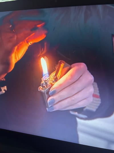 A close-up shot shows a person holding a lit golden lighter in their left hand. The hand holding the lighter has blue nail polish. The person is wearing a dark top with red and white stripes. The image appears to be taken from a video or screen. Location details are not discernible from the image.