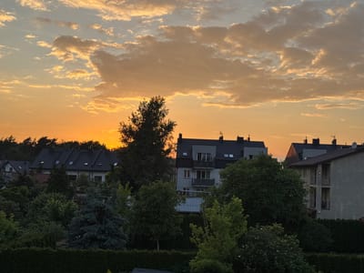The image depicts a residential area in Marki, Poland, during sunset. The sky is filled with warm hues of orange and yellow, with scattered, fluffy clouds. In the foreground, a dense arrangement of trees and evergreen bushes creates a natural barrier. Behind the greenery, several multi-story buildings with dark sloped roofs are visible. Some of these buildings feature balconies and visible windows, indicating residential use. The overall scene conveys a peaceful, suburban atmosphere at dusk.