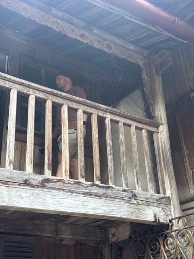 A low-angle shot reveals a weathered wooden structure, possibly part of a building, in Sà Phìn Commune, Vietnam. A man is visible behind a ladder-like series of horizontal wooden slats; his head is visible on the left side of the slats and his torso is obscured in the slats. A sign with a no smoking symbol is affixed to the wall to the right of the structure. The surroundings appear to be made of aged wood with intricate carvings.