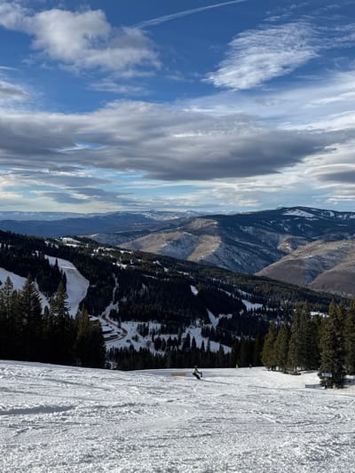 A landscape of snow-covered mountains and sky dominates the scene. The sky features blue patches interspersed with elongated white and gray clouds. Mountains stretch across the middle ground, exhibiting varying shades of brown and green interspersed with snow. A ski slope covered in textured snow fills the foreground, with a lone skier visible. Trees dot the mountainside, creating a contrast between snow and vegetation.