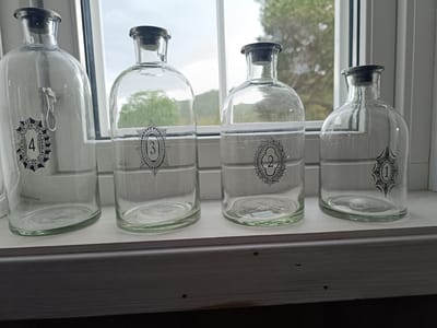 Four empty glass bottles are lined up on a windowsill. Each bottle has a black stopper and a decorative black label with a number in the center. From left to right, the numbers are 4, 3, 2, and 1. The first bottle has a small metal latch on its side. The window behind the bottles offers a view of trees and a cloudy sky, suggesting it is daytime and potentially overcast weather. The bottles are placed on a white wooden ledge, which is part of the windowsill. The setting appears to be indoors, with the window looking out onto an exterior landscape. There are no people visible in the image. The overall scene is still and domestic.