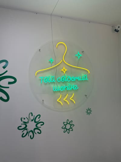 The image depicts a close-up, eye-level shot of a neon sign mounted on a white wall. The sign is circular with a yellow neon outline of a hanger at the top. Inside the hanger, the words "Твій свій одяг" are written in vibrant teal neon. Below this, in yellow neon, are three stylized chevron shapes pointing downwards. The sign is illuminated by neon lights, casting a glow on the wall. To the left of the sign, a large green stylized swirl or flourish is partially visible. Several smaller green circular embellishments resembling stylized flowers or snowflakes are scattered on the wall around the main sign. A security camera is visible in the upper right corner of the image. The lighting suggests an indoor environment. The image location is identified as Sarny, Ukraine.
