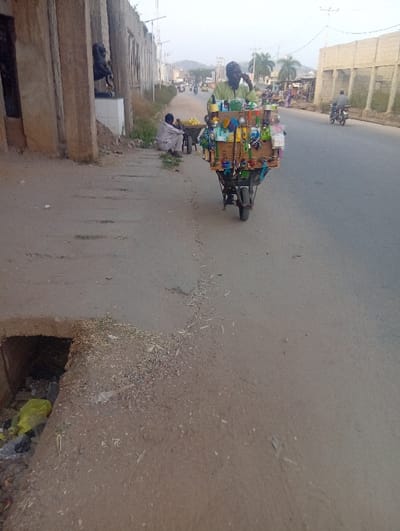 The image depicts an everyday street scene at Itakpe Junction, Nigeria, likely during the early morning or late afternoon due to the soft, diffused natural light. The central figure is a male street vendor, dressed in a light green long-sleeved shirt and a dark cap, riding a custom-built, three-wheeled motorized cart. He is engaged in conversation on a mobile phone, held to his ear.

His cart functions as a mobile shop, densely packed with a diverse array of goods. Visible items include sponges, brushes, various plastic containers, packaged products, and colorful small trinkets or jewelry, indicative of a general convenience store on wheels. To the left of the main vendor, closer to the foreground, another man is seated on the dusty unpaved shoulder of the road. He is wearing a light-colored tunic and is positioned next to a wheelbarrow filled with yellow fruits, possibly lemons or oranges.

The road surface is paved but appears worn, and the shoulder is unpaved and somewhat littered. In the immediate foreground on the left, an open drainage ditch or culvert contains some trash, including a distinctive yellow plastic bag. Along the left side of the street, there are concrete buildings, some of which seem unfinished or weathered. One structure features a dark statue, possibly depicting an animal or mythological figure, set upon a white pedestal. On the right side of the road, a concrete block wall runs parallel to the street. In the background, the road stretches into the distance, with other vehicles, primarily motorcycles, and hazy hills under a pale sky. There is no visible text in the image. The scene captures typical roadside commerce and daily life in a Nigerian locale.