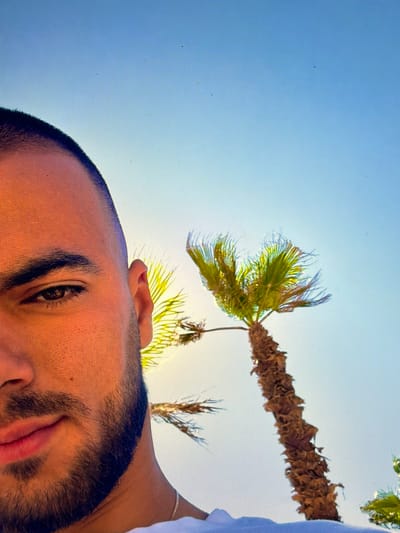 The image presents a rotated, low-angle perspective, predominantly featuring a man's partial face and a palm tree against a clear blue sky. The man's face occupies the left and bottom portions of the frame (when viewed in its current orientation), with his right eye, eyebrow, forehead, nose, and mouth visible. He has short dark hair and a dark, full beard or heavy stubble. His right eye is open, looking upwards, and his lips are slightly parted. A portion of his right ear and shoulder, covered by a white garment, are also visible, along with a delicate silver chain or bracelet on his right wrist/arm.
Above the man, against a bright, uniform light blue sky, the top of a palm tree is featured. Its rugged brown trunk, appearing to be covered in dried husks, extends diagonally from the upper right, with vibrant green fronds fanning out from its top, catching the sunlight. Other, less defined palm fronds are also hinted at in the background. The bright sky and strong lighting suggest a sunny daytime setting with clear weather. The scene conveys an outdoor, warm climate, consistent with the location context of Rosen, Bulgaria, if it implies a coastal or resort area where palm trees are found. There is no visible text in the image.