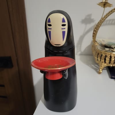 A black figure with a pale mask featuring purple triangles for eyes and mouth stands on a white surface. The figure holds a small red dish with a gold rim. To the right of the figure is a decorative gold object. In the background, a portion of a dark brown vertical surface is visible to the left, and a white wall occupies the rest of the background. The image does not provide any information about the city of Глебовский, Russia.