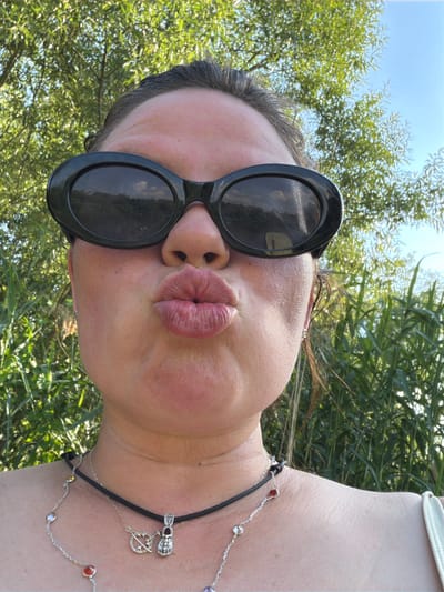 This is a portrait of a person outdoors, likely in Kamianka-Buzka, Ukraine, as suggested by the context. The subject is wearing large, black, oval-shaped sunglasses and has their lips pursed into a kiss. Their head is tilted back, and the image is shot from a low angle, with the sky visible above and lush green foliage in the background. The natural light suggests it is daytime, likely sunny. The person is also wearing a necklace with various charms and colorful beads. The overall mood appears playful and candid.