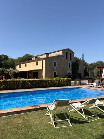 This image captures a tranquil scene at a rural property in Bescanó, Spain, featuring a swimming pool and a rustic stone house. The bright blue swimming pool dominates the foreground, its surface rippling under the clear blue sky. On the edge of the pool, a small orange and black paddleball racket rests near the tiled edge. In the grass beyond the pool, three white folding lounge chairs are arranged, two of them angled slightly towards the viewer, with one chair on the far right partially visible.

Behind the pool and chairs, a wooden fence adorned with dense, yellow flowering bushes separates the pool area from the property's main building. The house itself is a charming, two-story structure with a light yellow stucco exterior on the left side and a rough stone facade on the right. It has a tiled roof and features several windows, some with arched tops. A covered patio area extends from the left side of the house, with tables and chairs visible underneath. Trees and greenery surround the property, creating a lush, natural setting. In the background, a tall electricity pylon stands against the clear sky, a hint of modern infrastructure in the otherwise bucolic landscape. A few people can be seen near the covered patio area on the left side of the house, appearing to be interacting casually. The overall impression is one of a peaceful retreat, perfect for relaxation under the Spanish sun.