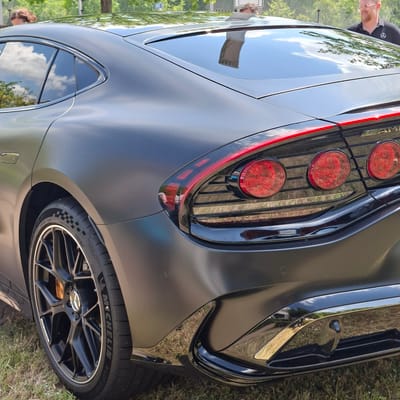 The image shows the rear quarter of a dark gray, matte finish sedan with a sleek, modern design. The car features prominent, round red taillights with intricate internal detailing, a thin red light strip across the trunk, and a black, glossy diffuser at the bottom. The car is parked on grass, with trees and foliage visible in the background. A man with glasses and a black polo shirt with a Mercedes-Benz logo is partially visible on the right, and another person's head and shoulder are visible on the far left, peeking over the car's roof. The reflection on the car's body suggests an outdoor setting, possibly a parking lot or event space. The overall scene is well-lit, indicating daytime. The question of location in Rastatt, Germany, is not directly supported by visual cues within the image itself.