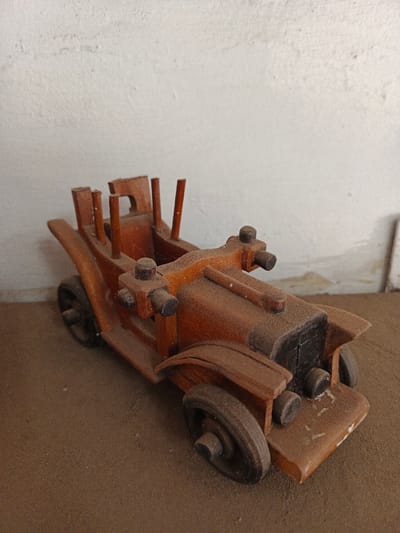 A dusty, vintage wooden toy car sits on a flat surface near a wall. The car is detailed, featuring a distinct front grill, fenders, wheels, and passenger compartment. Its wooden construction is evident, with a warm, aged hue. Dust coats the entire surface, muting its color. The background is a plain, textured wall, adding a simple backdrop to the scene.
