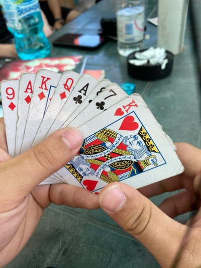 In an outdoor setting, likely in Gorna Malina, Bulgaria, a person is holding a fan of playing cards. The cards visible include a 9 of Diamonds, an Ace of Diamonds, a King of Diamonds, a Queen of Diamonds, an Ace of Clubs, a 7 of Clubs, and a King of Hearts. The King of Hearts is particularly prominent, with the full illustration of the king visible. The cards appear slightly worn. In the background, out of focus, are a blue plastic water bottle, a black ashtray with crumpled white tissues, and other people and objects on a green table. The lighting suggests it is daytime.