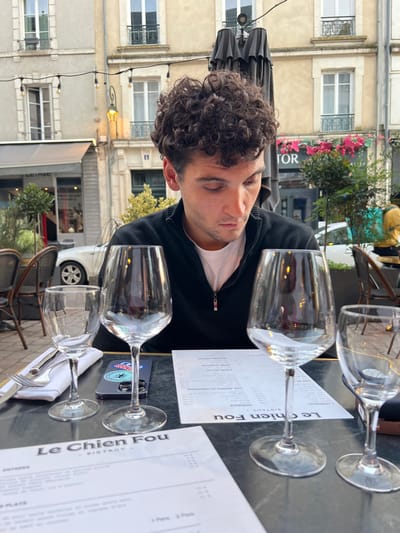 A young adult male, characterized by dark, curly hair and wearing a dark quarter-zip sweater over a white t-shirt, is seated at an outdoor bistro table with a dark surface. He is looking down intently at the table. On the table are four empty stemmed glasses: one smaller water or wine glass, two standard wine glasses, and a fourth partially visible wine glass. Two menus are present; the one in the foreground clearly displays "Le Chien Fou Bistrot." A dark-colored smartphone, featuring a palm tree sticker, is positioned between two of the glasses. Silverware, consisting of a fork and knife, rests on a white napkin to the left. The background features a European street scene with light-colored stone buildings, one of which displays the street number "13." Another building shows a partially visible sign reading "TOR" adorned with pink floral arrangements. String lights with exposed bulbs are strung overhead. Additional elements include dark outdoor cafe chairs, a streetlamp, a white sedan parked in the distance, and green potted plants. This scene is located at Le Chien Fou Bistrot, 2 Rue de la Cerche, 45000 Orléans, France.