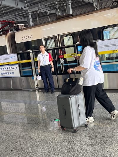 A woman in a white t-shirt and black pants walks with a silver suitcase and a black bag on wheels in a train station in Tiaodeng, China. She is wearing white sneakers. A second woman, in a white uniform shirt and dark pants, stands to her left, holding a white cone-shaped object and looking at the first woman. Both women are on a tiled platform next to a train. The train has the number "050111" on its side. There are signs with Chinese text on the platform and on the train doors. The environment appears to be indoors, with metal beams and overhead structures visible.
