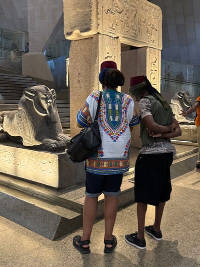 In Kafrat Nassar, Egypt, two individuals, viewed from the back, stand near an ancient Egyptian monument. The person on the left wears a white, colorful dashiki, dark denim shorts, and dark sandals. They carry a black messenger bag over their left shoulder. The person on the right wears a light-colored, patterned shirt, a dark green vest, black shorts, and black sneakers. Both individuals wear red fez hats. They are positioned in front of a stone structure with hieroglyphics, which appears to be part of an ancient Egyptian exhibit, possibly in a museum. A stone sphinx statue is visible to the left of the monument. The environment is indoors, with modern architectural elements like glass railings and escalators visible in the background, suggesting a museum setting.