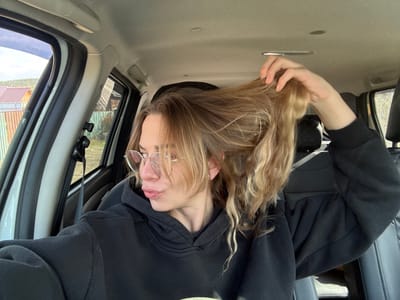 A woman is inside a car, upside down. She has blonde hair, is wearing glasses, and a black hooded sweatshirt. She is holding her hair. The car interior is light-colored. Outside the car window, there is a view of buildings and a hillside, suggesting an outdoor location. The city of Ust'-Ilimsk, Russia, is not discernible in the image.