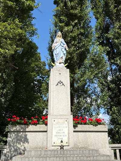 This image depicts a statue of the Virgin Mary atop a granite monument in Ostrzeszów, Poland. The statue, dressed in a blue and white robe, stands against a backdrop of lush green trees and a bright blue sky. The monument itself is adorned with red flowers in planters and features an inscription in Polish. The inscription reads: "NAJSLODSZE SERCE MARII BADZ NASZYM RATUNKIEM 1946" which translates to "Sweetest Heart of Mary, be our rescue 1946". The monument is made of grey granite with some decorative elements, including an emblem that appears to be two crossed rifles. The scene is set outdoors, suggesting a public space or memorial.