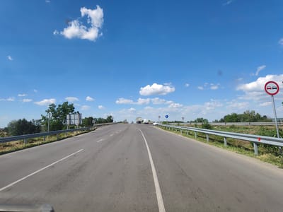 The image shows a highway in the city of Yasen, Bulgaria, on a clear day with scattered clouds. The perspective is from a vehicle traveling on the road. The highway has two lanes in each direction, separated by a white line. Guardrails line both sides of the road. In the distance, several vehicles are visible, including cars, a truck, and a van. Traffic signs are present, including a sign indicating "no overtaking" and other road signs. The surrounding environment consists of trees and greenery along the roadside. The sky is blue with fluffy white clouds.