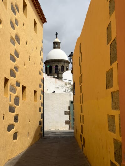 A narrow, paved alleyway in Agüimes, Spain, is presented, flanked by two multi-story buildings. The building on the left exhibits an ochre-yellow stucco facade with embedded dark grey, irregularly shaped stone blocks and several small, rectangular recessed openings. A tiled roof edge is visible at its apex. The building on the right also features an ochre-yellow facade with dark grey rectangular stone inserts arranged vertically. The alley floor is shadowed. Further down the alley, a white wall with dark stone accents is visible, accompanied by a small green door. A black bird is perched on the upper edge of this white wall, near sparse green foliage. In the background, under an overcast grey sky, a white domed church structure with a dark cupola and a cross is prominent. Its lower section reveals arched windows and grey stonework. Multi-colored pennants are strung across the church's facade. A dark, ornate street lamp is partially visible beside the dome. No individuals are present in the scene.