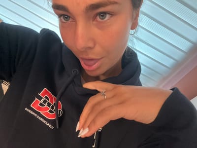 A person is posing for a selfie, likely indoors. The person is wearing a black hooded sweatshirt with the logo "Danderyd Djursholm Elite" printed on the chest. The person has light eyes, fair skin, and dark eyebrows. They are wearing a ring on one of their fingers and have manicured nails with a white design. Behind them are horizontal blinds. The photo provides no information about the city of Kashin, Bulgaria.