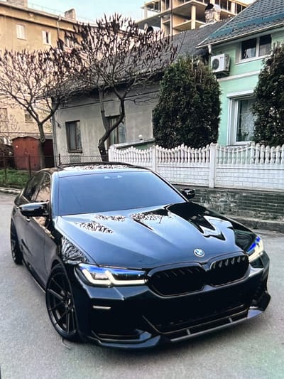 An angled, eye-level, front-view shot captures a black BMW sedan parked on a residential street. The car's sleek design is highlighted by its glossy finish and the blue accents in its headlights. Behind the car, a light green house with a dark green roof stands adorned with a white picket fence and manicured evergreen shrubs. Further in the background, a multi-story building under construction, with its exposed concrete framework, contrasts with the established residential structures. Bare trees with twiggy branches frame the scene, adding a touch of nature to the urban setting. The street is paved with asphalt, and the sky above is overcast, suggesting a cool or cloudy day. No people are visible in the frame. The overall impression is one of a quiet, suburban street.