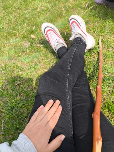 A person wearing black jeans and white athletic shoes with red accents is sitting on green grass, with their legs crossed. A long, thin wooden stick lies parallel to their right leg. The person's left hand rests on their left thigh, fingers splayed. The environment is outdoors with visible blades of grass and sunlight casting shadows. The image was captured in Anastasivka, Ukraine.