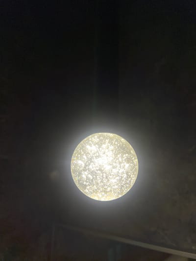 A single, illuminated, orb-shaped light fixture is the central subject of this image, set against a dark, ambiguous background. The orb is textured with numerous small, irregular bubbles or inclusions, which catch and refract the light, giving it a sparkling, ethereal quality. The light emanating from the fixture is bright and appears to be a warm, white tone, casting a soft glow that illuminates the immediate surroundings. The background is predominantly black, with subtle variations in shade suggesting depth or texture, possibly a ceiling or a wall. There are faint linear elements visible in the upper right portion of the frame, which might indicate structural features of the setting. The overall impression is one of a dimly lit environment, possibly indoors at night, where the light fixture is the primary source of illumination. There is no indication of people or specific activities taking place. The visual focus is on the abstract beauty and texture of the illuminated orb. No text is visible in the image.