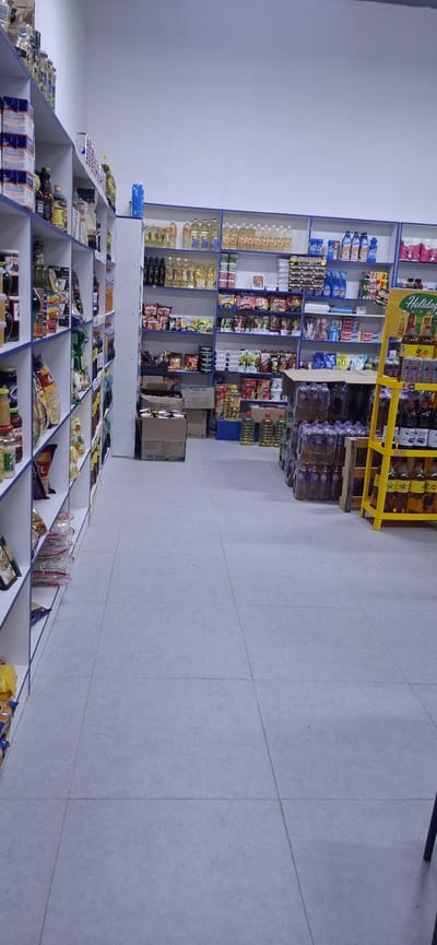 The image shows the interior of a small grocery store or market located in Arys, Kazakhstan. The store features white shelving units filled with various food products and household items. On the left, shelves are stocked with packaged goods, bottles, and jars. On the right, larger shelves display bottles of cooking oil, cleaning supplies, and other packaged items. A yellow display stand is visible in the foreground, holding bottles of drinks. The floor is tiled in a light grey color. The overall environment is organized and well-lit, typical of a retail setting. There are no people visible in the image.