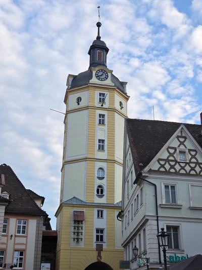 A tall, pale yellow tower with a dark gray, conical roof and a weather vane stands against a partly cloudy sky. The tower has a clock on one side and several rows of bells mounted on its facade. To the right of the tower is a light blue building with a dark gray, tiled roof and decorative patterns on its gables. To the left of the tower is a building with a pale pink and white facade, also with a dark gray, tiled roof. The scene appears to be an urban setting in Ansbach, Germany, likely a town square or street, given the presence of multiple buildings and architectural styles.