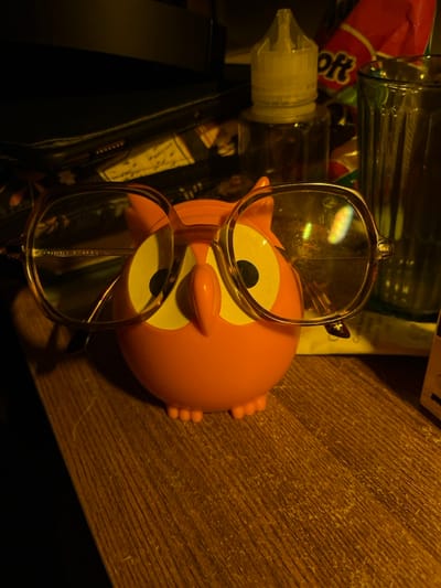 The image shows an owl-shaped object with glasses resting on it. The owl is orange and faces the camera with large, white eyes. Behind the owl, there is a wood-like surface. Other items in the image include a bottle with a dropper, a glass jar, and a red and pink package. The overall lighting is dim, giving the image a warm tone.