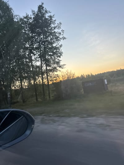 The image depicts a roadside scene in Orlivka, Ukraine, likely captured from a moving vehicle. The sky is a pale blue, transitioning to a warm, golden hue near the horizon, indicating either sunrise or sunset. Several trees with dark green foliage are visible on the left, their branches silhouetted against the bright sky. In the background, a dark, rectangular structure with white text is situated amidst a wooded area. The text on the structure appears to read "CULIAC." The ground to the right is a gray, textured surface, possibly asphalt, with a blurred appearance suggesting motion. The lower right corner of the image shows a portion of a dark, rounded object, which is likely a side-view mirror of the vehicle. The overall mood is serene, with the soft light of dawn or dusk casting a peaceful glow over the landscape. The presence of the trees and the distant structure suggest a rural or semi-rural setting.