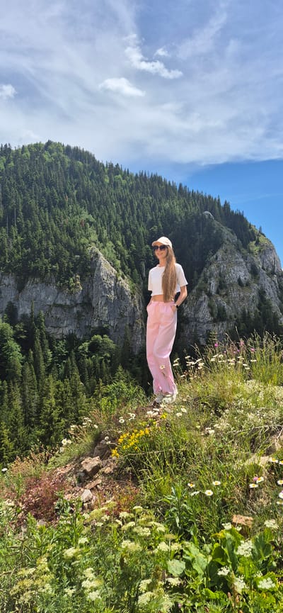 A young woman stands on a grassy, flower-strewn slope overlooking a steep, forested mountainside. The woman, facing the viewer, wears a white crop top, pink and white striped pants, a beige baseball cap, and sunglasses. Her long, blonde hair extends down her back. She has her hands in her pockets. The mountainside behind her is covered in dense evergreen trees, with rocky outcrops visible. The sky above is partly cloudy with patches of blue. The location is Sedlačka gora, Montenegro.