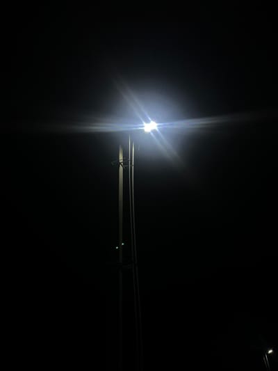 A street light illuminates a dark sky, casting a bright glare that extends across the upper portion of the image. The light source appears to be at the top of a tall, slender pole that is partially visible on the right side of the frame. Two green lights are visible on the pole, positioned below the main light. The background is entirely black, indicating nighttime and a lack of surrounding artificial or natural illumination, typical of a rural or less developed area. A second, fainter light is visible in the far lower right corner, suggesting another light source, possibly another street light or a distant building light. The scene is devoid of any people or active movement. The setting is consistent with a street in Бучацінскі сельскі Савет, Belarus, at night.