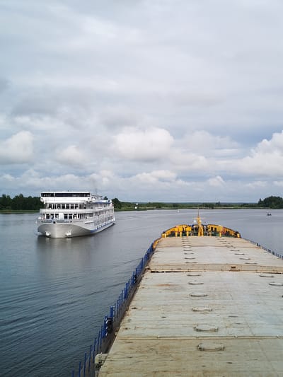 This is a vertical, eye-level, medium shot taken from the deck of a barge, looking forward towards a large white passenger ship sailing on a wide, calm body of water. The barge in the foreground has a long, flat, weathered deck that takes up the right two-thirds of the frame. The deck is composed of large, rectangular panels with raised, circular or oval hatches scattered across its surface. The far end of the barge is visible, showing yellow structures and blue railings. The passenger ship is on the left side of the frame, heading towards the viewer and slightly to the right. It is a multi-decked vessel with many windows and a white superstructure. The ship is emitting a thin plume of smoke from a smokestack. The water is a deep blue-grey, with faint ripples. In the background, a distant shoreline is lined with dense green trees, and the sky is overcast with large, dramatic grey and white clouds. A small, distant boat is visible on the water to the right. The image appears to be taken in a river or canal, as indicated by the presence of navigation markers and the relatively narrow waterway. The location is identified as Kargulino, Russia.