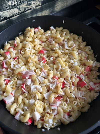 A close-up, overhead shot shows a black pan filled with macaroni pasta and imitation crab meat. The pasta is cooked and tossed with a creamy dressing, and the imitation crab is shredded into small pieces, mixed throughout the dish. Some of the imitation crab pieces have a red outer layer, creating a visual contrast with the white pasta. The dish appears to be a prepared meal, possibly a pasta salad. The background shows a blurry patterned surface, possibly a tablecloth or placemat, with a dark, tiled backsplash visible above it. The location of Smolyaninovo, Russia is not discernible from the image.