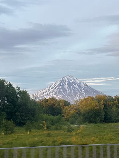 A snow-capped, conical mountain dominates the background of this image, set against a pale, cloudy sky. The mountain's slopes are streaked with dark rock and white snow. In the middle ground, a dense line of trees, mostly green with some autumn-colored foliage, forms a natural barrier. A few horizontal power lines traverse the sky above the trees. The foreground consists of a grassy field with a blurred metal railing at the very bottom, suggesting the photo was taken from a moving vehicle. The scene appears to be of a natural landscape, likely outside of urban development, and no people or specific structures are visible. The presence of a snow-capped mountain suggests a region with significant elevation or a colder climate. Without further context, it's impossible to definitively confirm the location as Кеткино, Russia, though the mountainous terrain is consistent with some Russian landscapes.