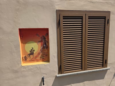 The image shows a section of an exterior wall in Viterbo, Italy. On the left side of the wall, there is a small, rectangular mural painted within a recessed orange-colored frame. The mural depicts a scene with a windmill, a large yellow sun, and two figures on horseback in silhouette against a gradient of orange and red. One horseman is larger and appears to be charging forward, while a smaller figure on a horse follows behind. The wall itself is painted a light beige color and shows signs of wear, including a crack running across the upper left quadrant of the image. To the right of the mural is a closed wooden shutter with horizontal louvers, installed within a brown frame. The shutter appears to be made of wood and is mounted on the wall. Below the shutter, there is a grey granite windowsill. A dark faucet or spigot is visible attached to the wall below the mural. The overall scene suggests an outdoor setting, likely on the side of a building in an Italian town.