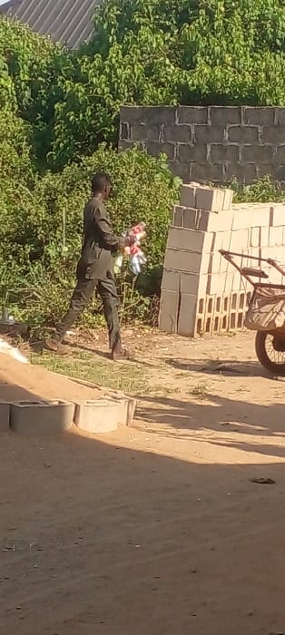 The image depicts an outdoor scene in Agunmo, Nigeria, captured during daylight hours. A single male individual, dressed in a dark-colored long-sleeved tunic and matching trousers, walks from left to right across a light-brown dirt path. He carries a bundle of multi-colored plastic bottles in his left arm.

In the midground, to the right of the individual, a stack of light-colored hollow concrete blocks is neatly arranged. Behind these blocks, the rear section of a single-wheeled handcart or wheelbarrow is partially visible, featuring a dark wheel and a beige, possibly fabric-covered, storage area. To the left of the path, in the foreground, several light-colored concrete elements, possibly curbing or block fragments, are present.

The background features dense green foliage and bushes, with a dark grey concrete block wall partially visible behind the stacked blocks. Above the foliage on the upper left, a dark corrugated metal roof is present. The ground is consistently dirt or dry earth.