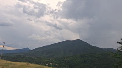 The image depicts a serene mountain landscape in Dobrotino, Bulgaria, under a dramatic, cloudy sky. The foreground is dominated by a grassy meadow, its golden hues suggesting either late afternoon light or the dry season. In the mid-ground, rolling hills covered in dense, dark green trees rise to meet a prominent, rounded mountain peak. The texture of the foliage is rich and varied, indicating a healthy forest. Scattered among the trees on the lower slopes are a few small houses with red roofs, hinting at a small rural settlement.

Further in the background, a series of layered mountain ranges fade into a hazy distance, their outlines softened by atmospheric perspective. A utility pole with wires stretches across the scene, connecting the present with the unseen. The sky is a tapestry of thick, grey clouds, with some patches of brighter white suggesting breaks in the overcast. A distinct column of darker cloud, possibly indicating rain or a storm cell, is visible rising behind the main mountain peak, adding a sense of dynamic weather to the scene. The overall mood is one of peaceful solitude, with the looming clouds adding a touch of dramatic natural beauty. The time of day is not clearly defined but likely late afternoon or early evening, given the quality of light on the meadow. There is no visible text in the image.