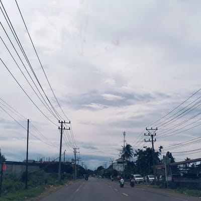The image depicts a busy road scene in Keutapang, Indonesia, under an overcast sky during daytime. The perspective is from the center of the paved road, looking forward into the distance.
**Subjects:** The prominent subjects are numerous overhead power lines and utility poles that stretch across both sides of the road, dominating the upper half of the frame. The road itself, marked with white lines, is a key feature. Various vehicles, primarily motorcycles and scooters, are visible on the road, with some heading towards the viewer and others in the distance. A few cars are parked or moving on the right side. Tropical vegetation, including palm trees, bushes, and other green foliage, lines both sides of the road. Several buildings, including what appears to be a construction site on the left and an open-sided structure possibly related to vehicle services on the right, are interspersed among the trees. A tall cell tower stands in the mid-right background.
**Activity or event:** The scene captures daily traffic and commuting activity along a local road. People are seen riding motorcycles, suggesting routine travel.
**Notable details:** The weather is characterized by a uniformly cloudy and overcast sky, indicating soft, diffused lighting with no direct sunlight or harsh shadows. The time of day is clearly daytime. The dense network of power lines and poles is a striking visual element. On the right side, there's a sign next to parked cars, but the text is unreadable. The overall atmosphere is that of everyday life in a semi-urban or rural area, with no strong emotional cues.