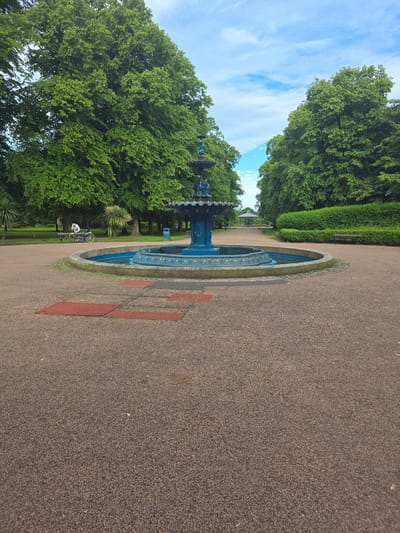 The image shows a wide view of a park, likely in Stockton-on-Tees, United Kingdom, dominated by lush green trees lining a gravel path. A prominent blue fountain is centrally located, with water features visible. In the middle ground, a person is seated on a park bench next to a bicycle, facing away from the camera. To the right of the fountain, a neatly trimmed hedge runs along the path, with a park bench visible in front of it. In the background, a covered structure and more benches suggest a recreational area. The sky above is partly cloudy with a bright blue hue. The foreground is filled with the gravel texture of the path, with a few scattered red rectangular markers.